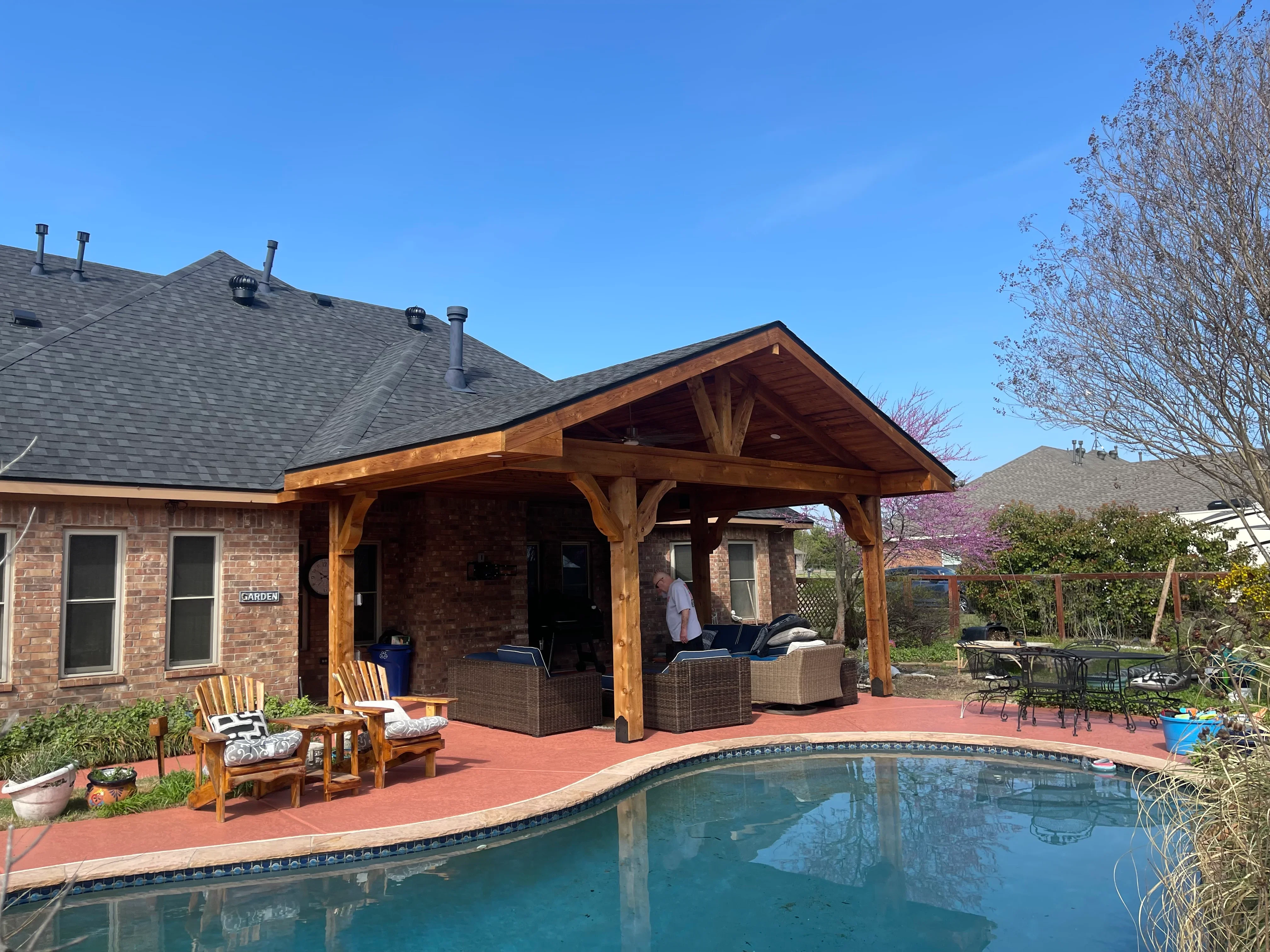 Wooden timber frame patio cover with gable roof attached to brick home, red tile patio, pool view