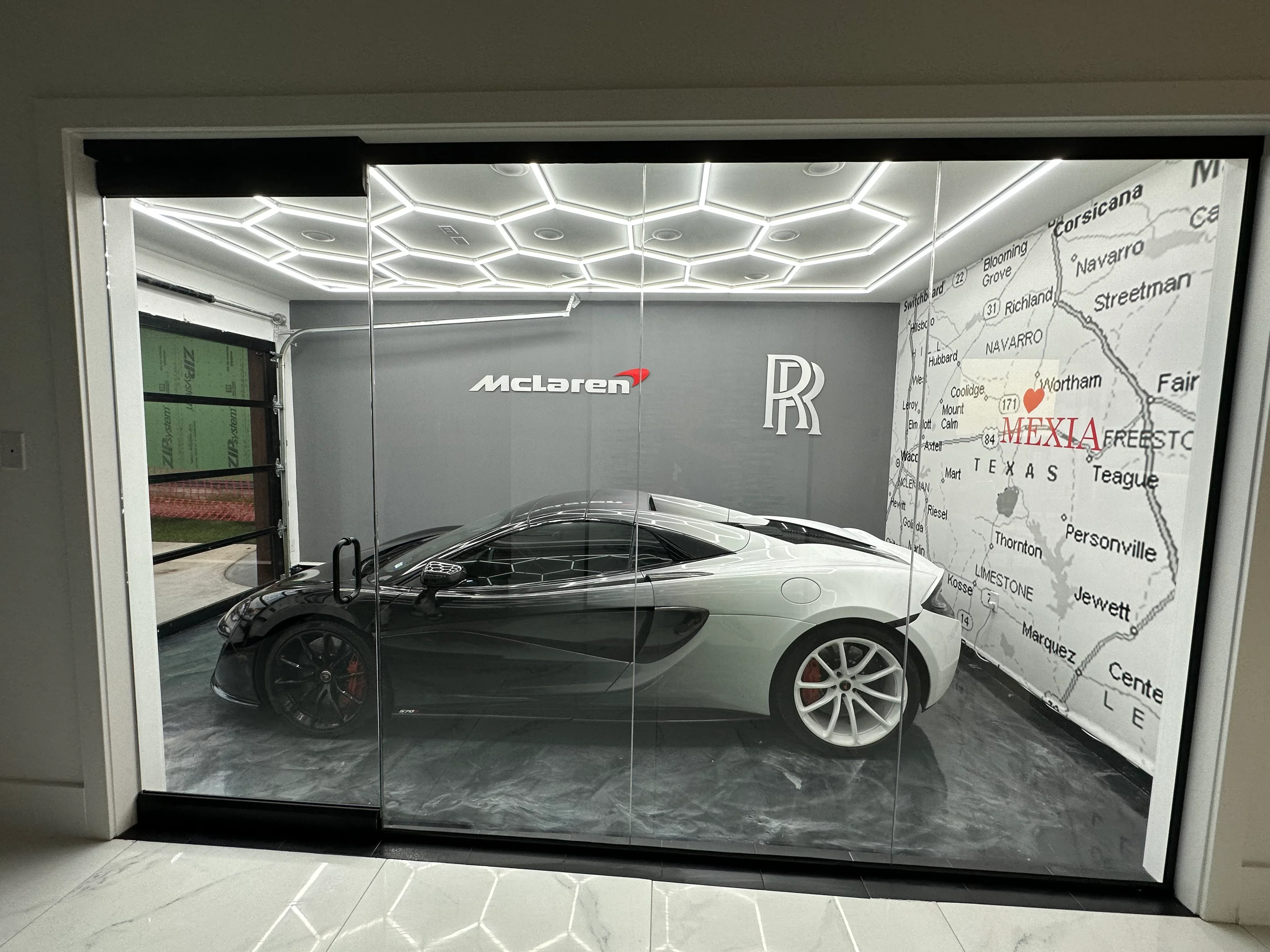 Luxury car showroom with white McLaren behind frameless glass, geometric LED ceiling, polished concrete floor